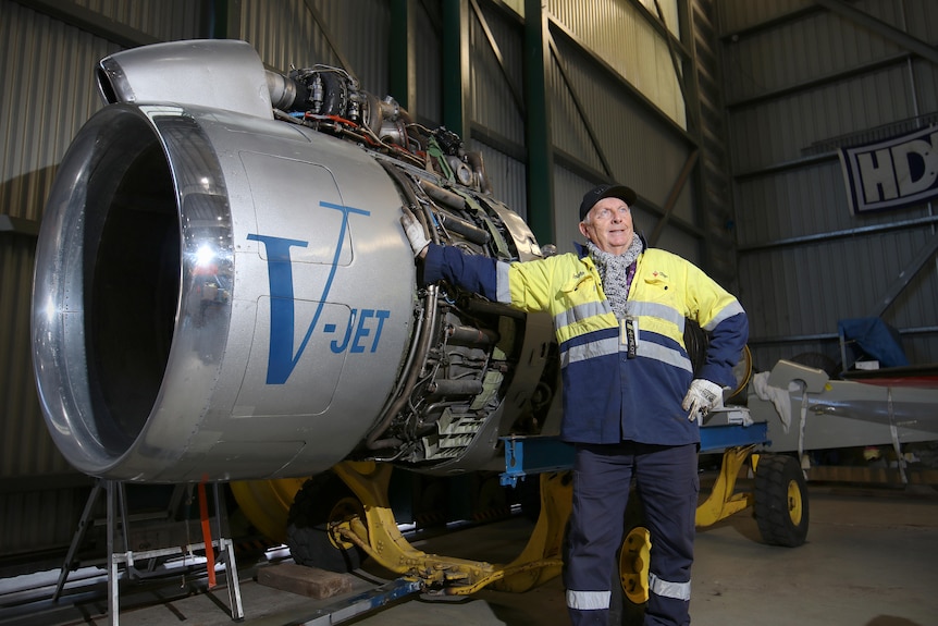 Bob De La Hunty leans against a large silver jet engine inside a hangar wearing a high vis shirt and gloves. 