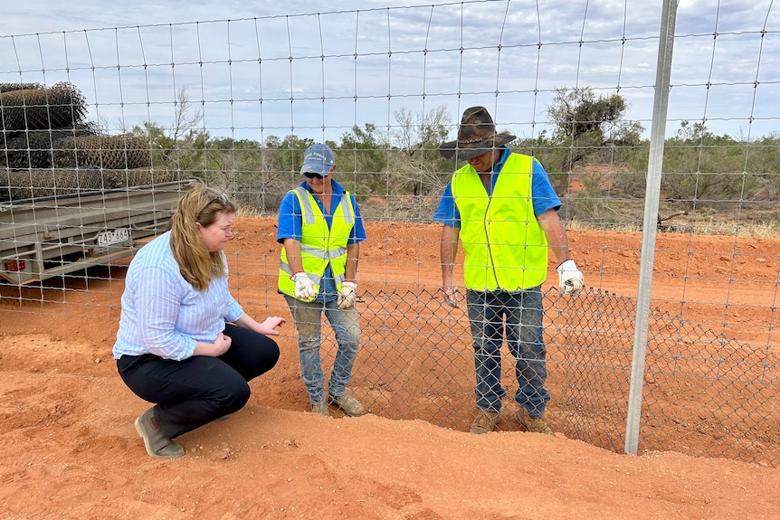 A women crouching down with two people in yellow vests standing along a fence line with red dirt and blue skys 