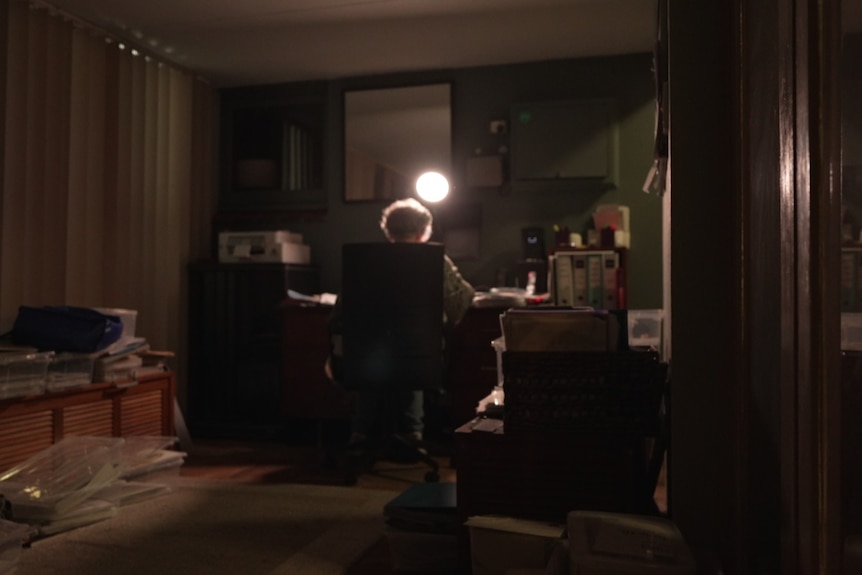 A woman with short hair is seated in a chair in a dark office, a singular light above her head. View is from the back of her
