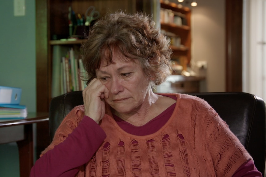 A middle-aged woman wearing tones of red in her top leans her hand on her cheek looking visibly upset