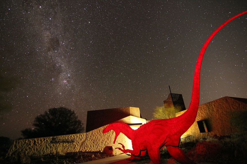A vividly-coloured mstatue of a dinosaur in front of a stone building beneath a spectacularly starry night in the outback.