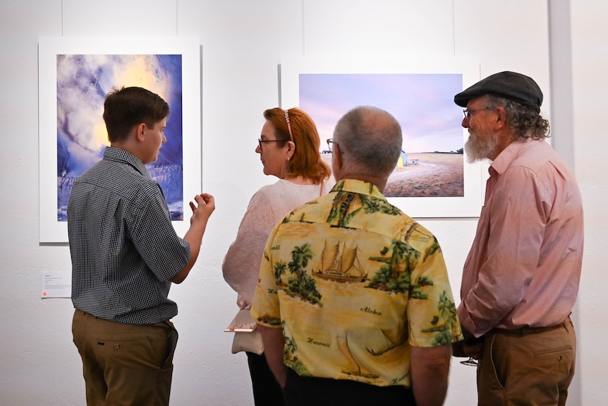 A young man speaks to a woman at an art gallery as two older men listen.