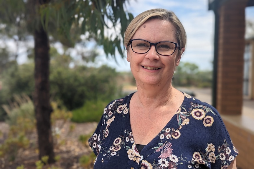 A woman with short blonde hair and glasses and a blue floral dress smiling at the camera.