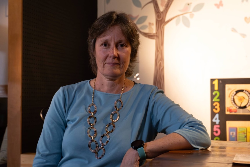 A woman with a long neckl;ace on and a blue top leaning against a set of drawers.