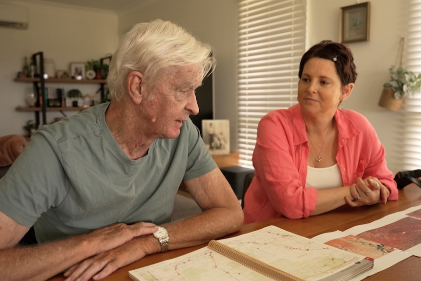 A man in his 80s and his adult daughter seated at a wooden kitchen table looking at each other chatting. Maps on the table