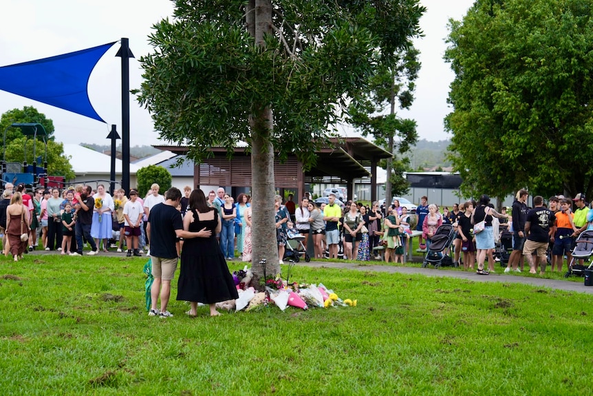 A crowd of people stand gathered in a park. Two people stand together next to a tree where people have laid flowers.
