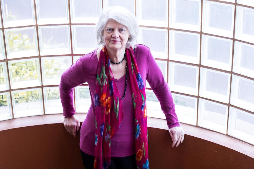 A woman with white hair and a pink cardigan stands news a window. 