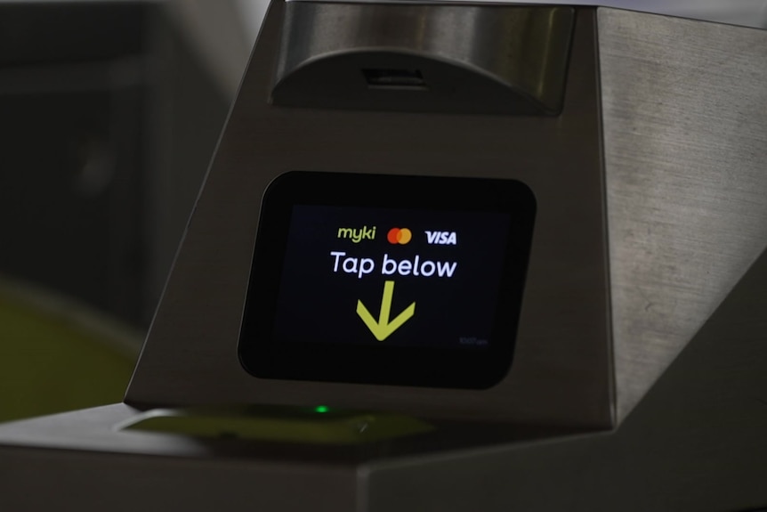 A silver machine with a black screen that says "myki", "visa" and "tap below" with a yellow arrow pointing downwards.
