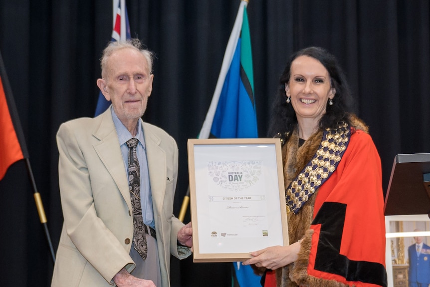 An elderly man holding a framed award with a woman wearing red robes.