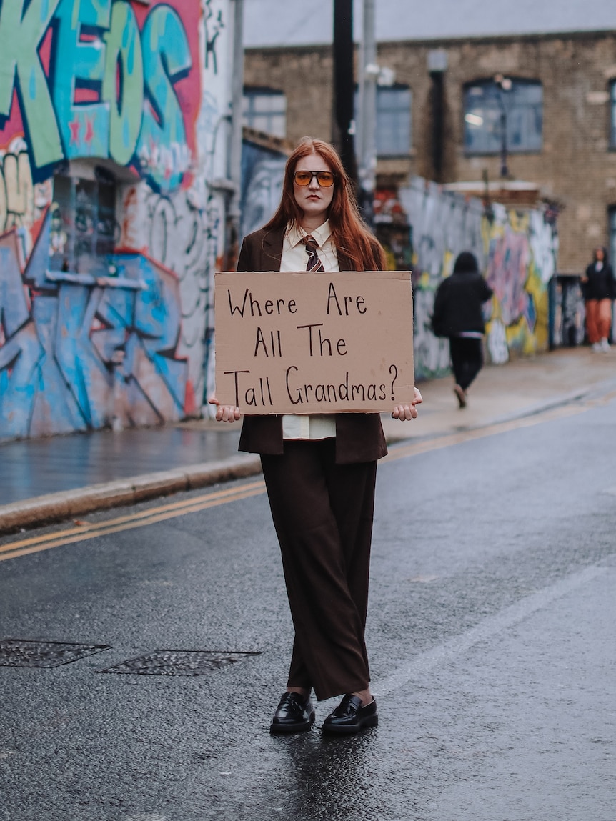 Lou Wall, late 20s, with long red hair, in 70s sunglasses and a brown suit, looks serious holding a cardboard sign on street.