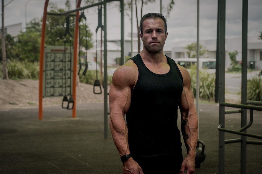 A man with muscled arms stands in a park, wearing a black tank top.
