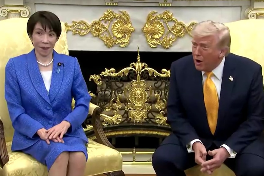 Japan's prime minister wearing a blue suit sits awkwardly with a surprised look as Donald Trump in a navy suit talks.