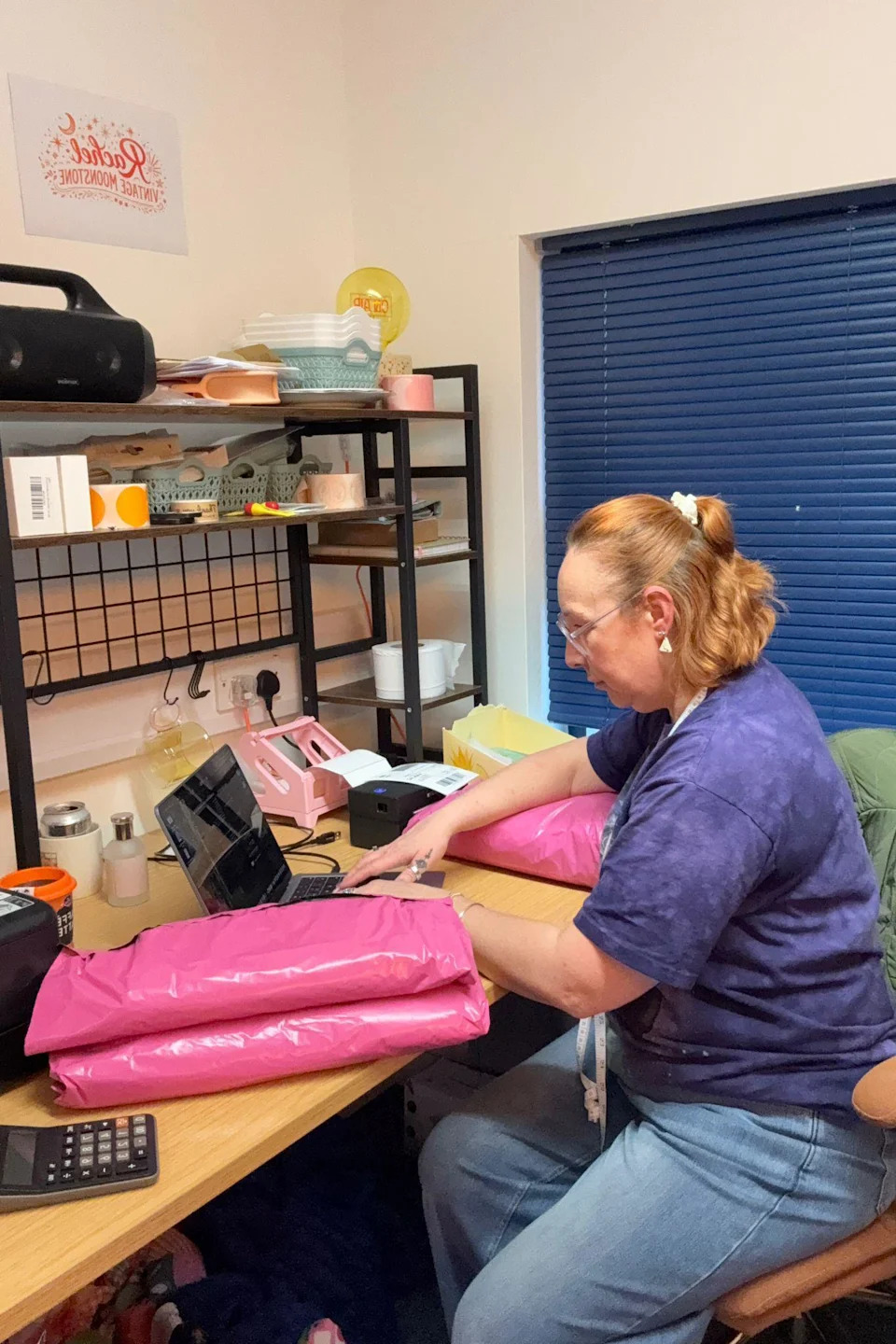 Rachel Grady sits at a laptop in front of a wooden desk. Two pink parcels can be seen on the desk, and she leans her right arm on another pink parcel. She has ginger short hair, which is half pulled up into a bobble and she looks down at the laptop. Blue shutters can be seen behind her.