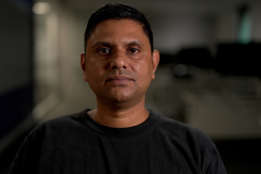 Man wearing a dark grey t-shirt sitting in a room with computers behind him.