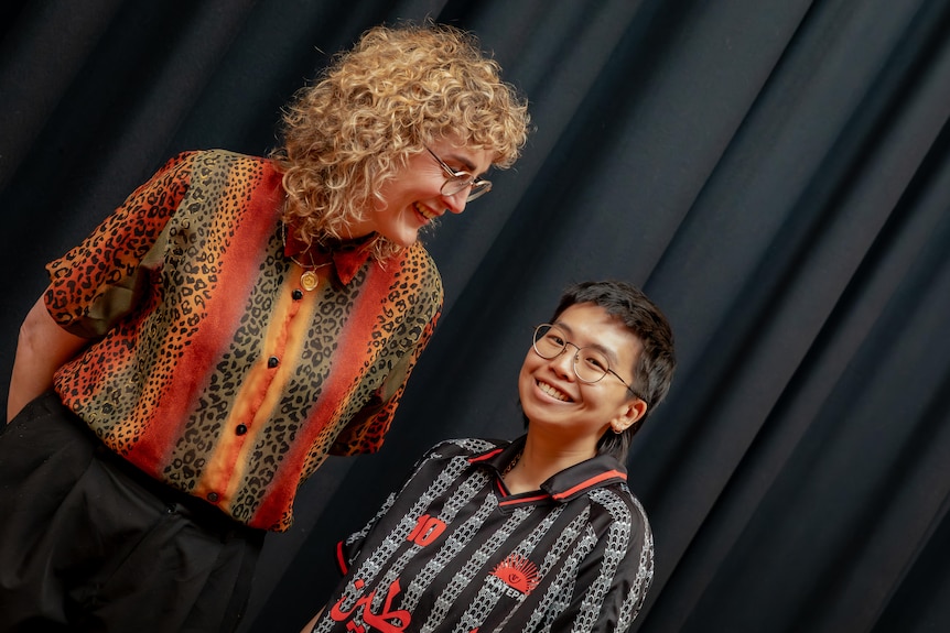 A tall blonde woman looks left at a shorter dark-haired person who is smiling, both in front of a black curtain