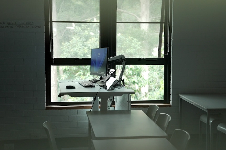 A dark classroom. A raised desk with a computer on it sits by a window. There are other tables and chairs.