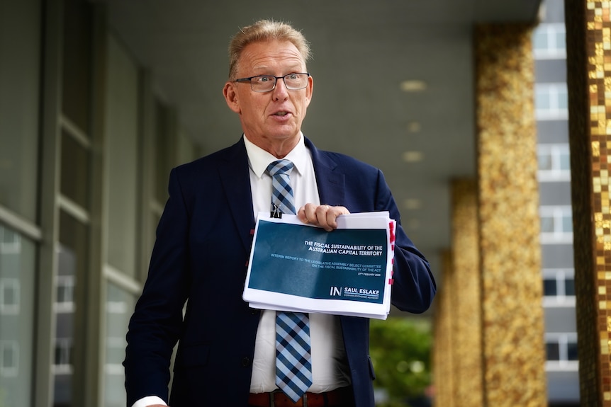 A man in a suit and tie holds up a bunch of budget papers