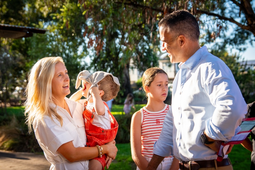 A woman holding a toddler and a man standing next to a young girl look at each other chatting