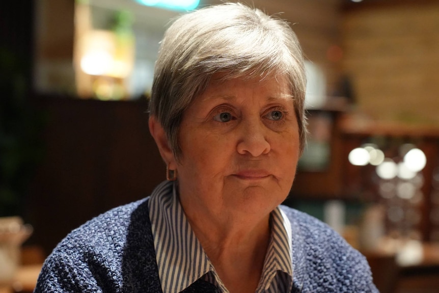 A grey haired woman wearing a blue cardigan and white striped button up shirt.