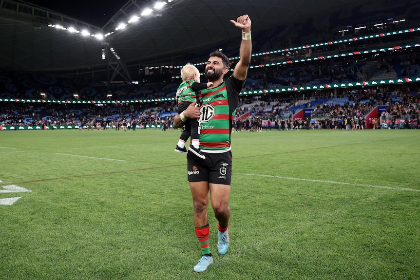 Alex Johnston waves to South Sydney Rabbitohs fans while holding his child after an NRL game.