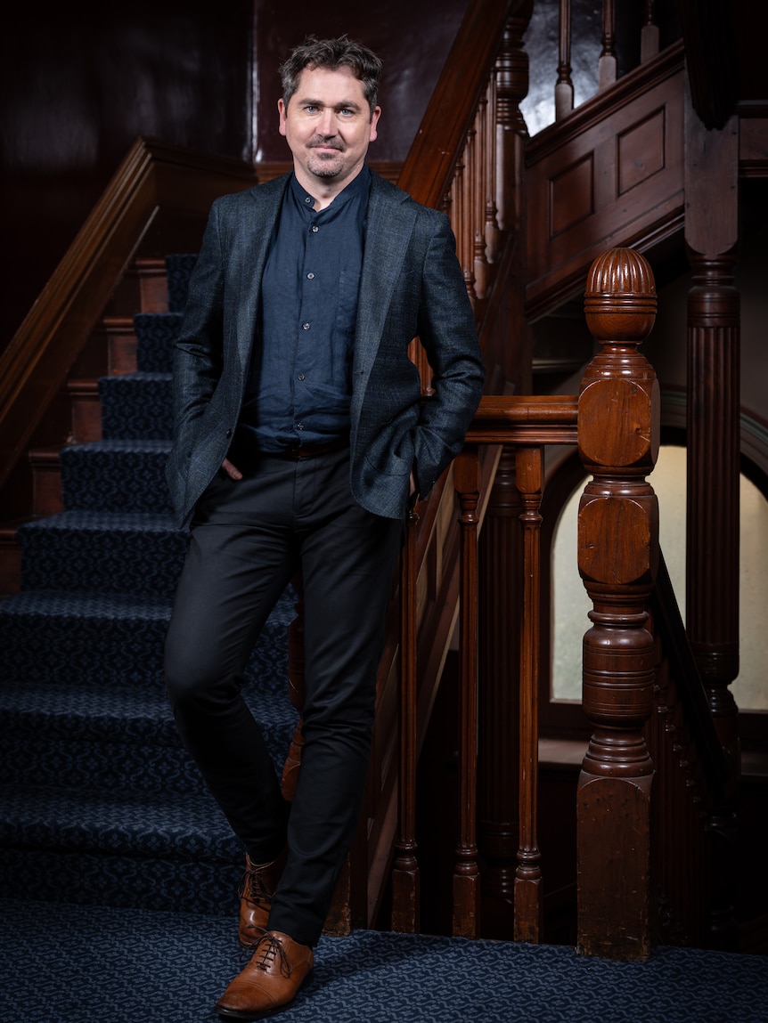 Matthew Lutton, 41, in a suit, smiles slightly, leaning back against a staircase balustrade.