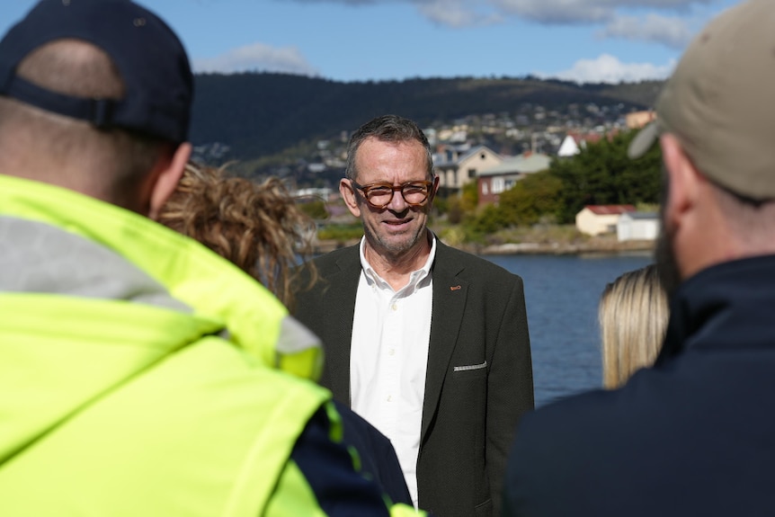 John Whittington in a suit and glasses stands on Battery Point foreshore with a group of workers in caps and hi-vis jackets.