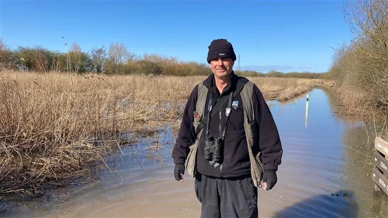 Pete is stood in a waterway.  He wears a gloves and a hat and carries a pair of binoculars around his neck. The reed bed is behind him.