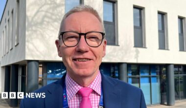 Andrew Morgan, who is looking into the camera and smiling. He is wearing a pink and white checked shirt, a pink tie, and a dark blue blazer. He has short, light blond hair and is wearing a pair of dark-framed glasses. Morgan is photographed outside a hospital building, which is a two-storey structure. The lower storey has dark grey pillars and floor-to-ceiling windows, whilst the upper floor is painted cream and has about eight, narrow, grey-framed windows visible.