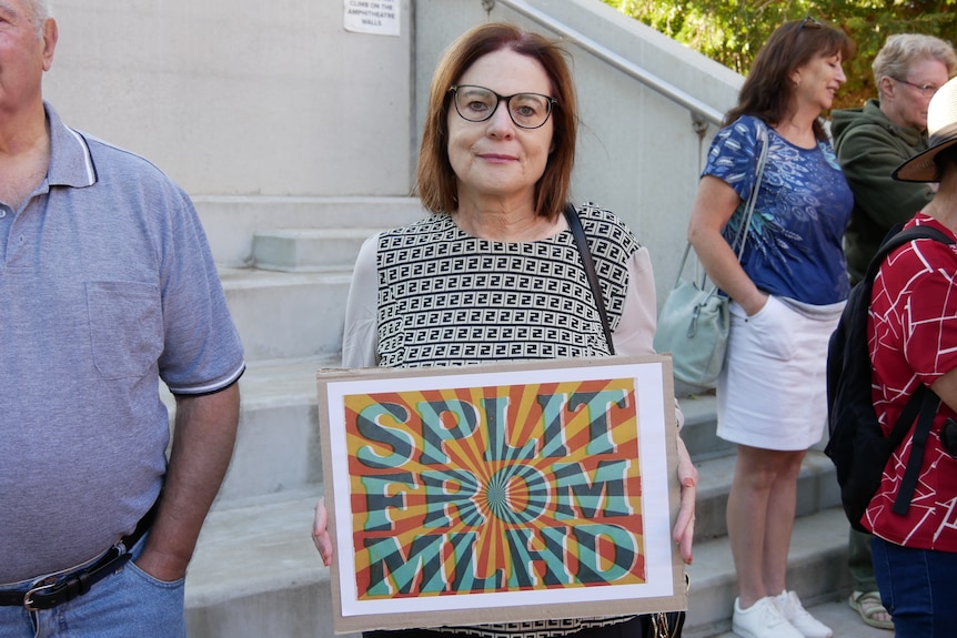 A woman with glasses stands with a colourful sign saying "Split From MLHD".