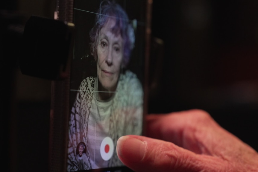 A mature woman with short hair that has coloured foils in front hovers her finger over the record button on an iPhone