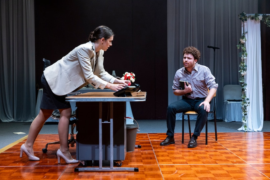 A stage; a woman wearing a blazer and high heels leans over a desk to speak to a man seated to her left.
