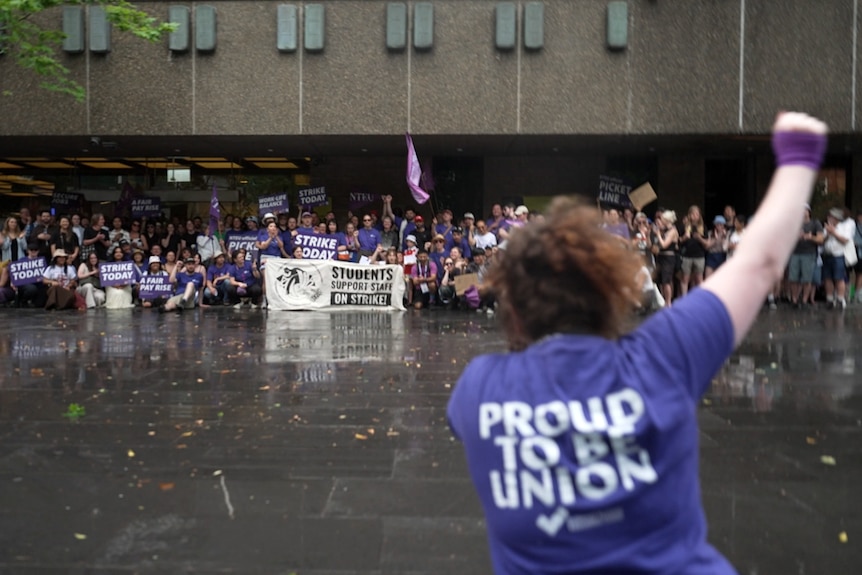 A person in a 'proud to be union' shirt holds up a fist. In front of them dozens of protesters stand in front of a uni building.