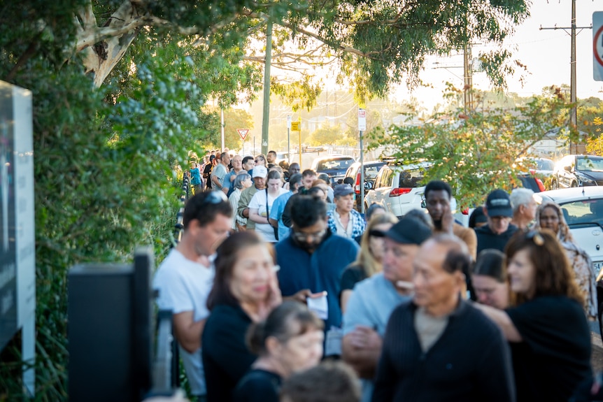 A long queue of people on a street with cars parked on one side