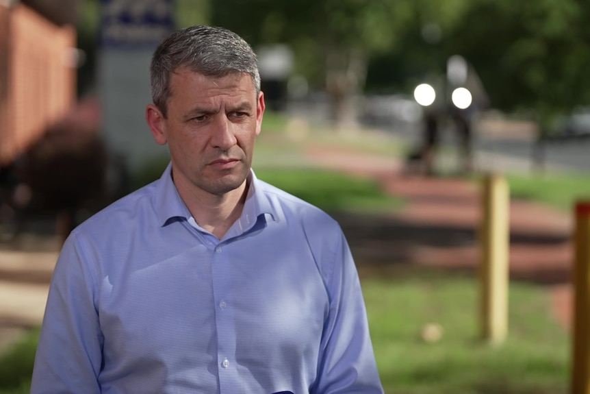 A man with short, grey hair wears a business shirt while standing on a grassed area near a footpath.