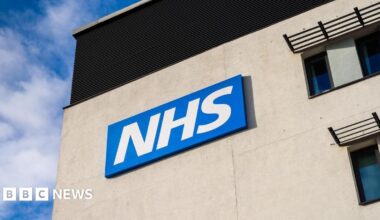 Bright white NHS lettering sits on a blue background, sitting high on the side of a grey building with blue sky visible behind it.