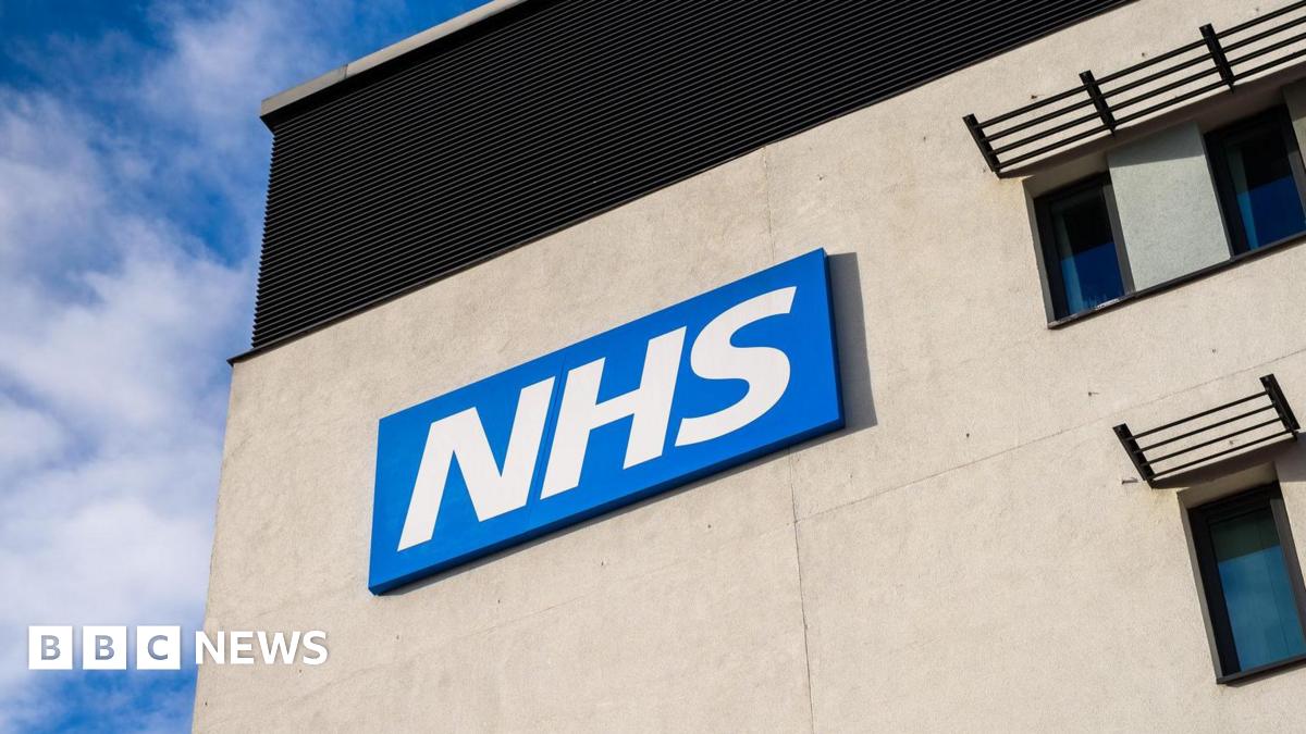 Bright white NHS lettering sits on a blue background, sitting high on the side of a grey building with blue sky visible behind it.