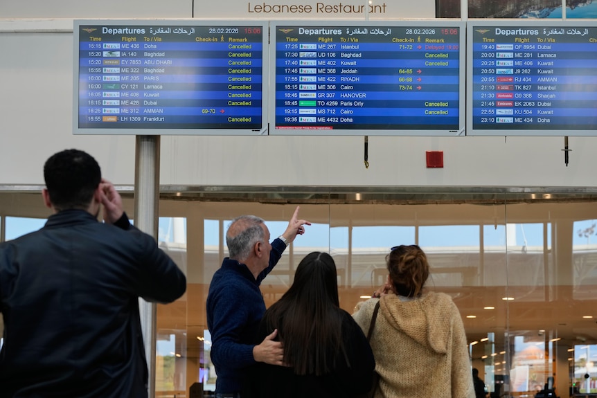 Four people stand and look at a departures screen at an airport. 
