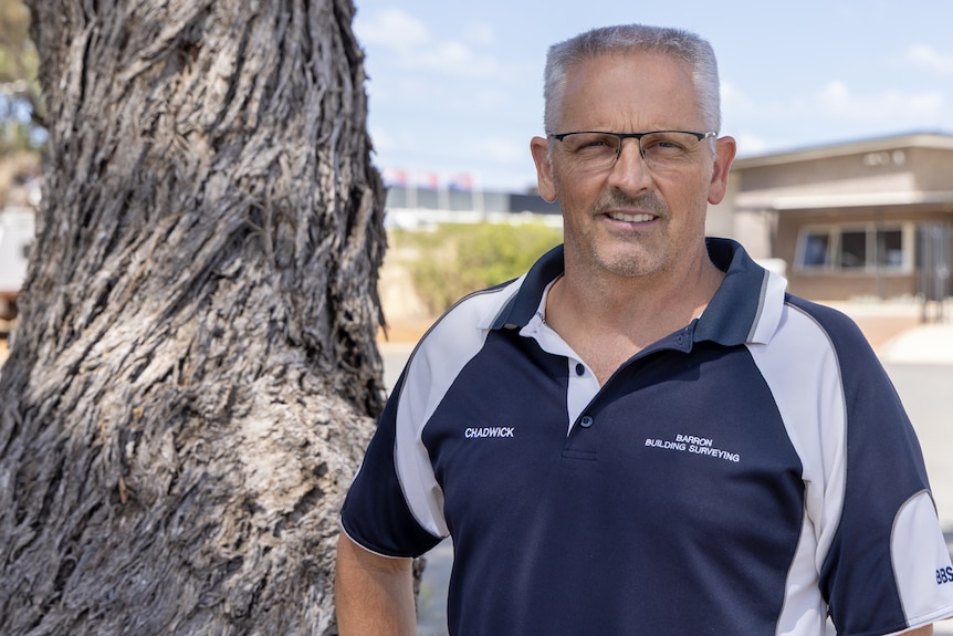 A man to right of camera with glasses and grey hair. Tree in background on left of camera
