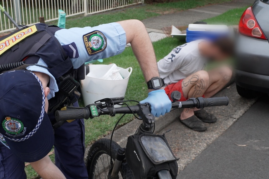 a police officer seizes an illegal e-bike as its owner sits on the ground in handcuffs