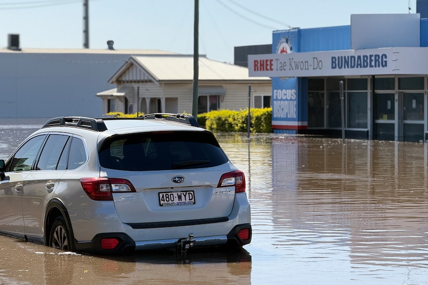 Flooded white car on street across from shopfront filled with brown floodwater