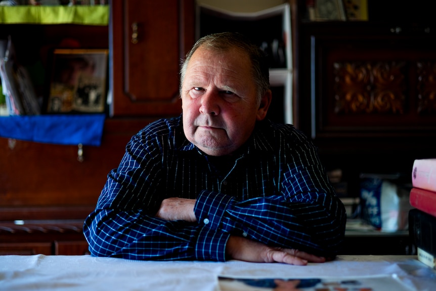 A man in a blue checked shirt has a pensive expression with crossed arms resting on a table