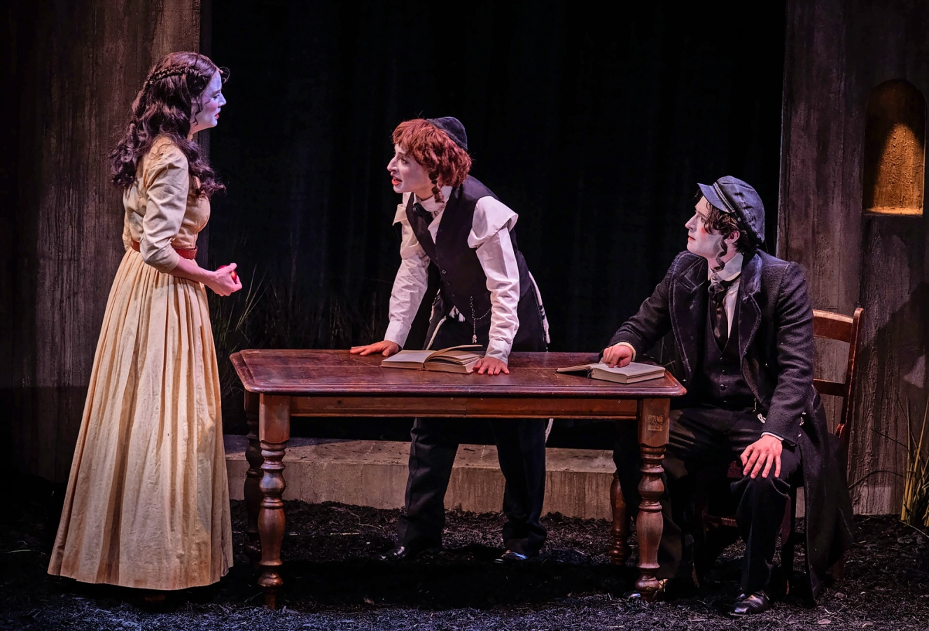 Three actors in costume on a stage, with a woman in a long dress facing two men dressed in traditional Jewish attire.