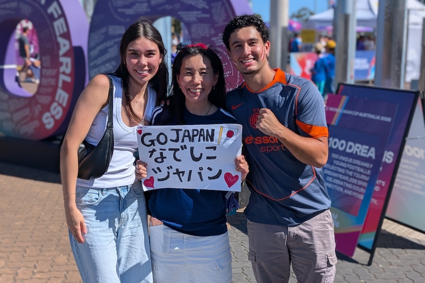 Three people stand and smile holding a Japan sign.