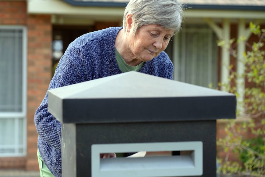 A grey-haired woman in a blue cardigan checking a letter box.