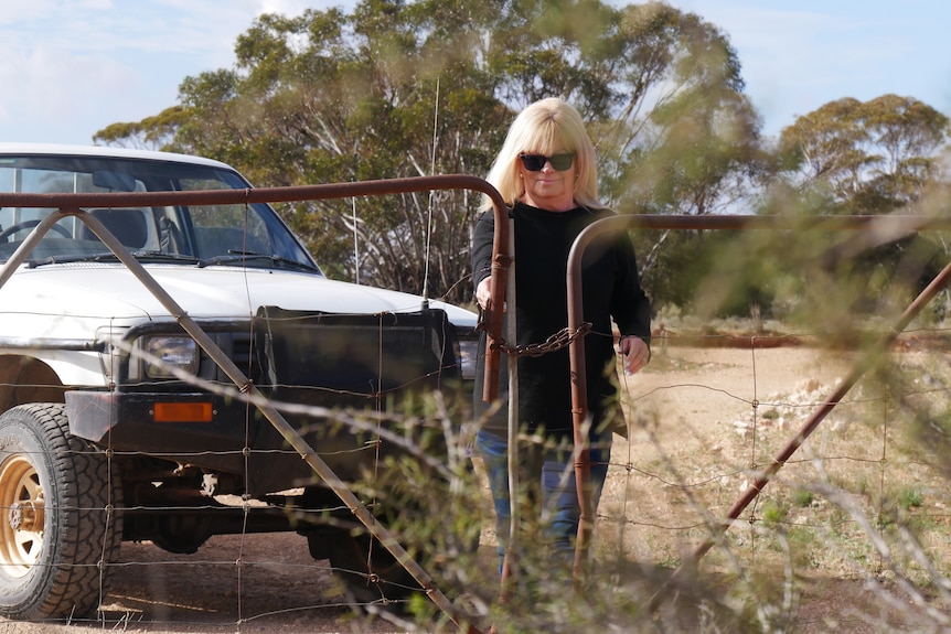 A woman opens a rusted gate in scrub land at her farming property.