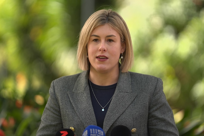A woman with chin length blonde hair in a grey jacket and black top stands in front of green trees.