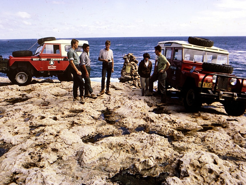 Two four-wheel-drive vehicles sit atop beachside rocks. six people stand around talking while standing next to vehicles