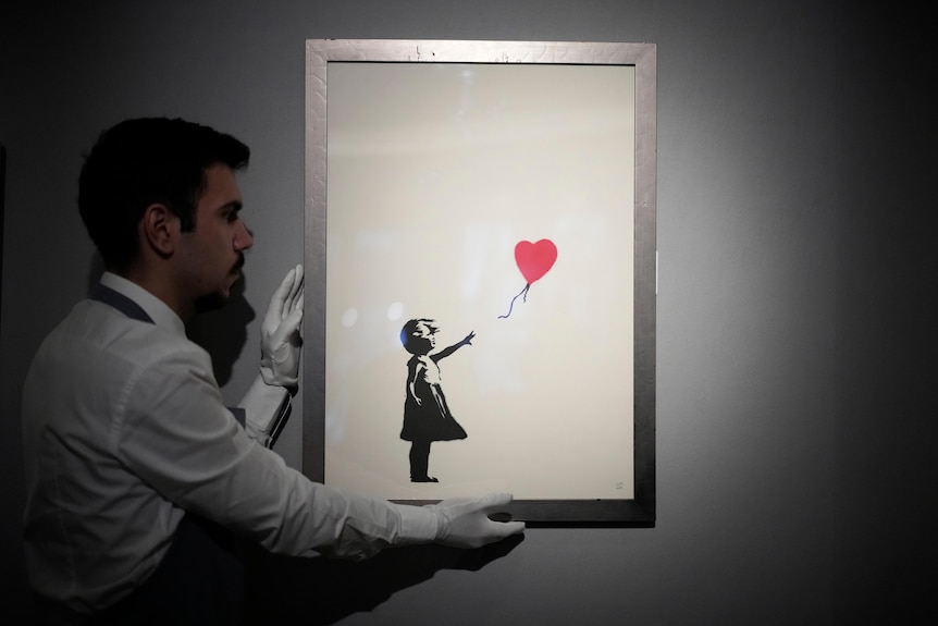 A man in a white shirt holds a framed image with white gloves; the image shows a girl and a heart-shaped balloon.
