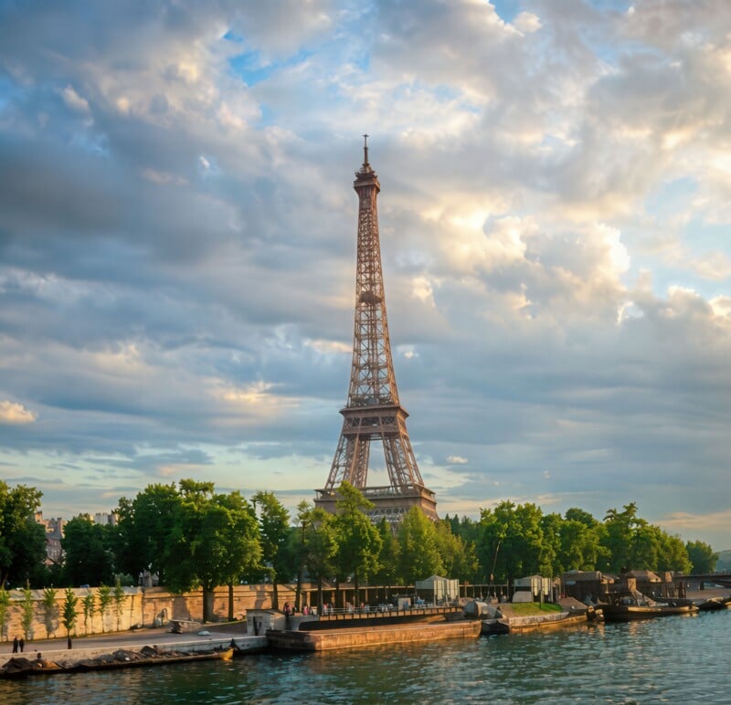 The Eiffel Tower rises above green trees along the Seine River in Paris, France, under a partly cloudy sky in soft, golden light.