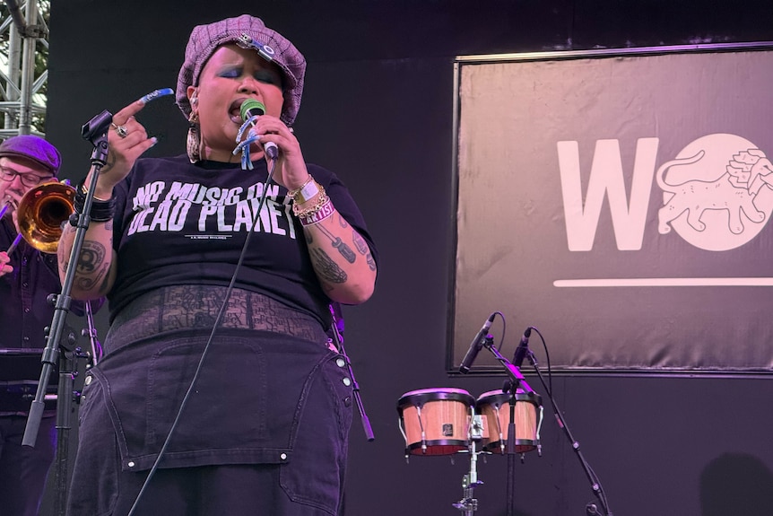 A vocalist onstage holding a microphone, wearing a slogan shirt, with percussion instruments beside them.